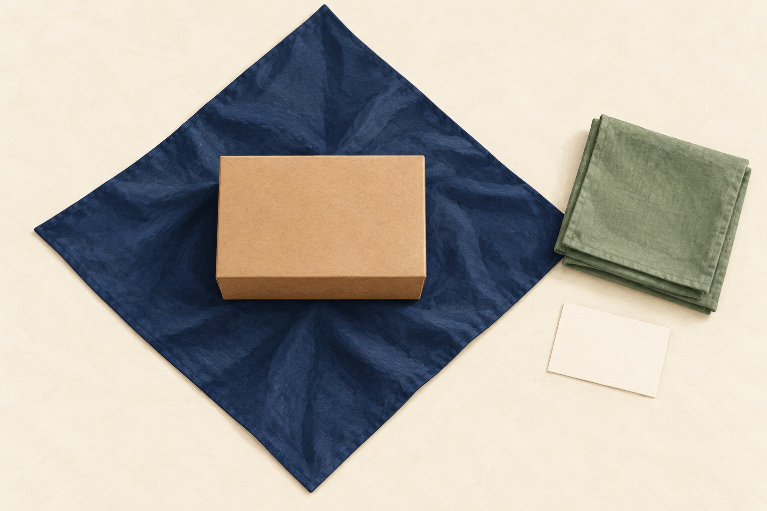 A closed kraft box rests on an open diamond-oriented navy cloth beside folded sage fabric and a blank card.
